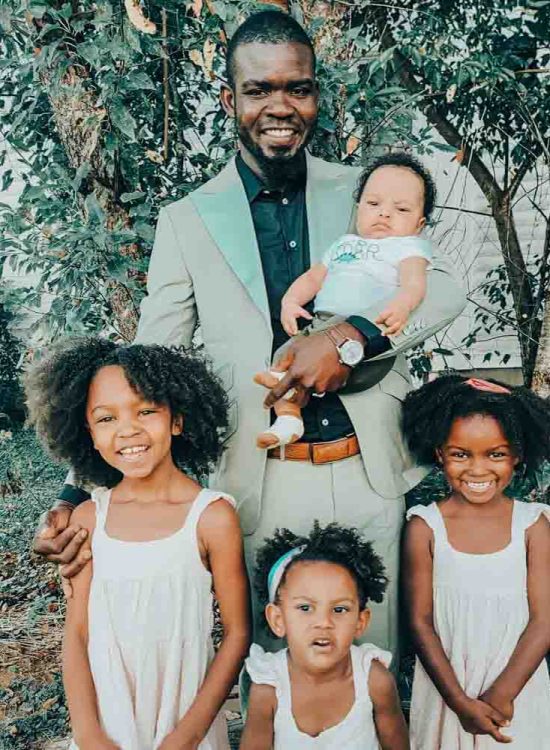 a man holding a baby and with his three kids standing on the backyard of their house