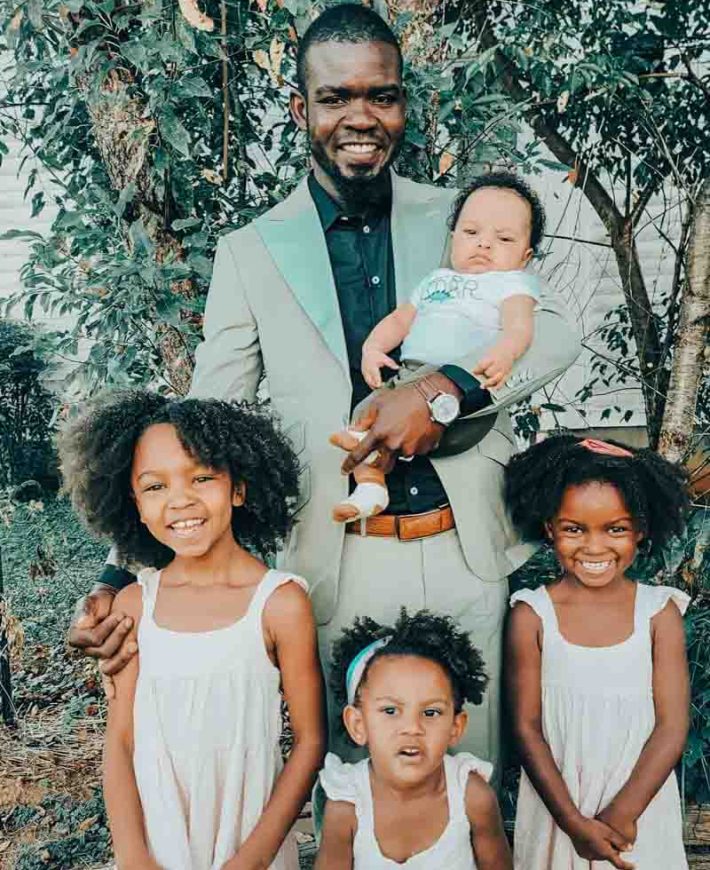 a man holding a baby and with his three kids standing on the backyard of their house
